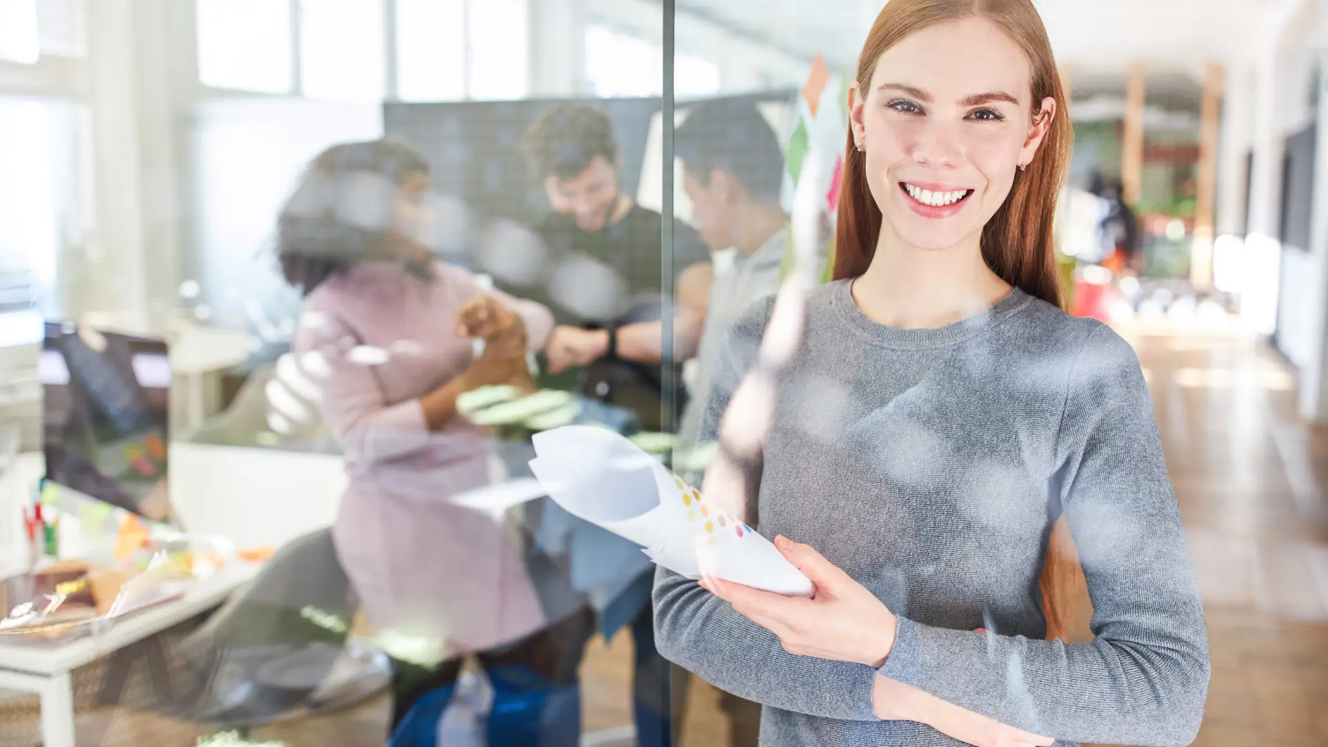 Smiling apprentice holding documents, with three more apprentices visible blurred in the background, talking to each other