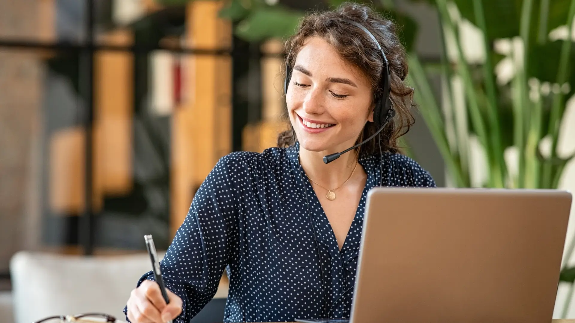 Friendly smiling woman with a headset in front of a laptop, writing something down with a ballpoint pen